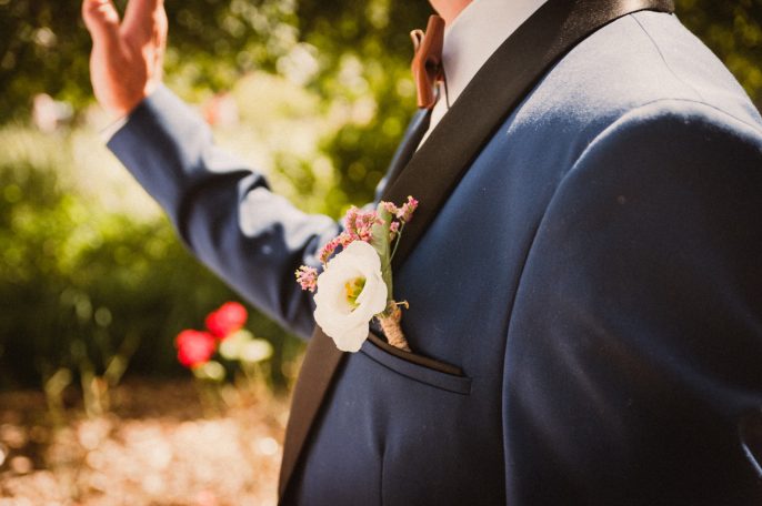 Homme en costume bleu avec une fleur dans la poche et un geste de la main.