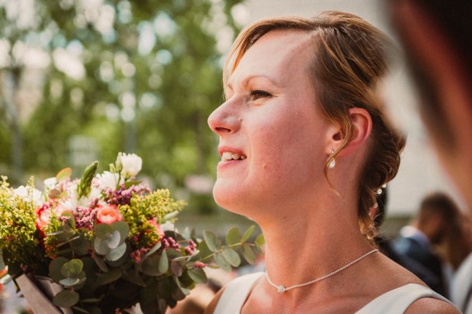 Femme souriante portant un bouquet de fleurs, en tenue de mariage, avec un fond flou.