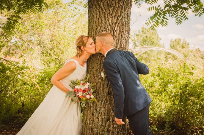 Un couple s'embrasse près d'un arbre, entouré de verdure et de lumière naturelle.