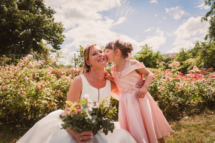 Femme en robe de mariée souriant à une petite fille lui donnant un bisou dans un jardin fleuri.